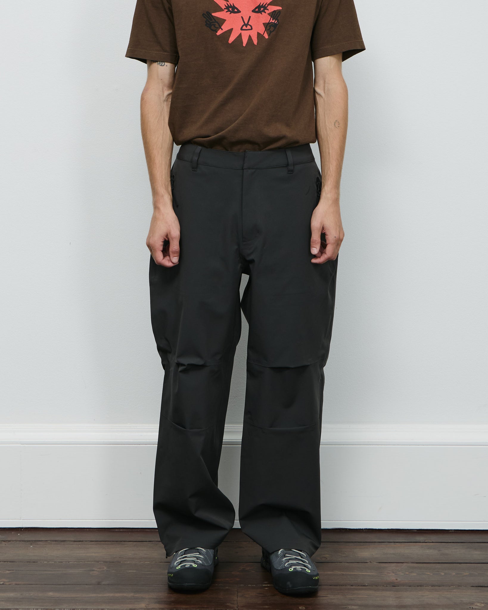 Person wearing a brown t-shirt with a graphic design and black pants against a white wall.